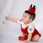 Cute baby wearing a red crown and outfit, reaching out playfully.