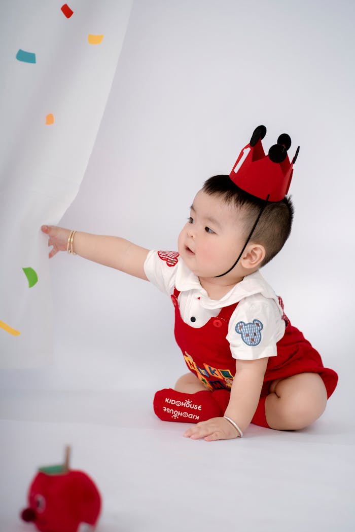 Cute baby wearing a red crown and outfit, reaching out playfully.