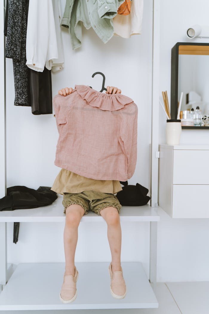 A child sits on a closet shelf holding a pink blouse playfully covering their face.