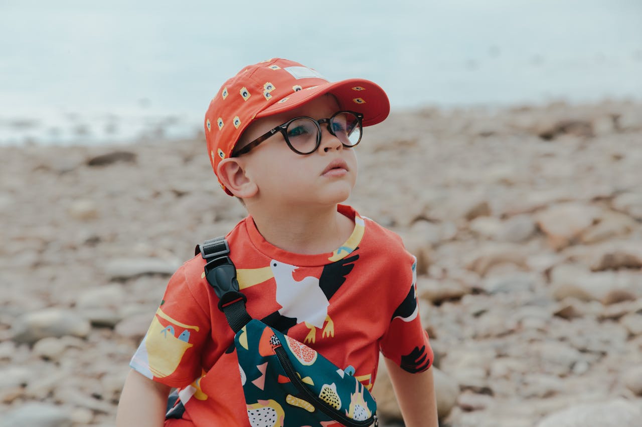 A child wearing a colorful cap and shirt with glasses, sitting on a rocky beach.